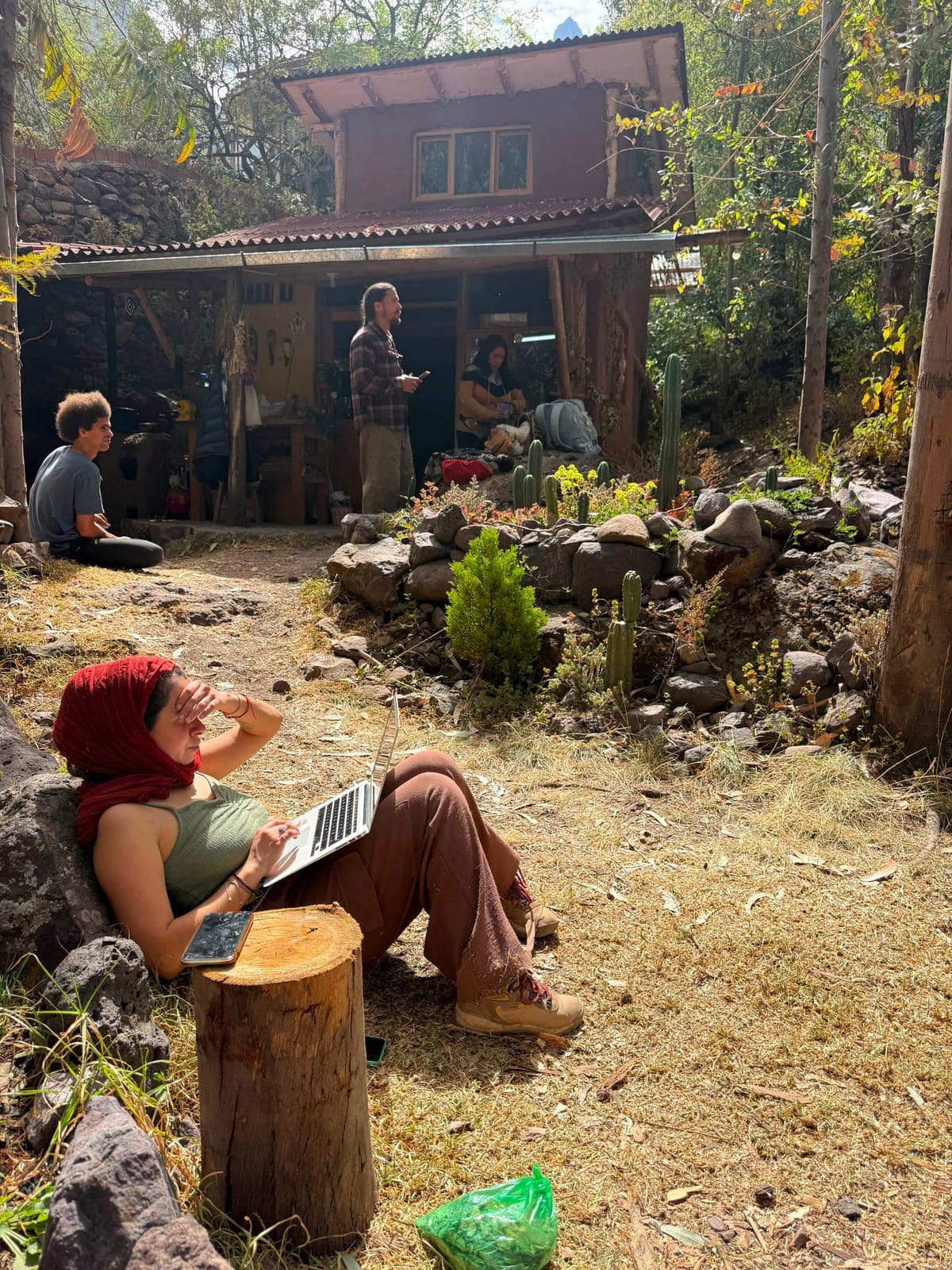 People gathered around a wooden cabin and garden before the ceremony, one woman reclining against a rock with a laptop