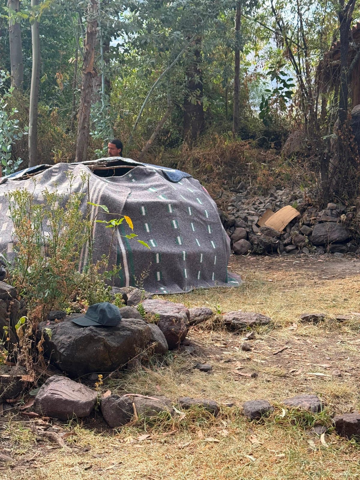 The finished sweat lodge covered in heavy grey blankets, a person's head emerging from the low doorway, a hat resting on a stone outside