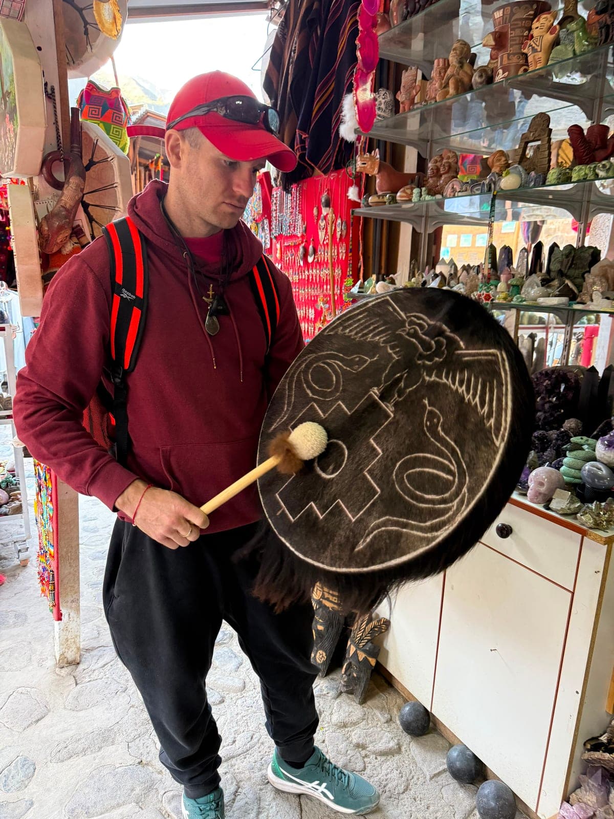 A man in a red cap playing a large hand-painted ceremonial drum inside an artisan shop lined with crystals and crafts