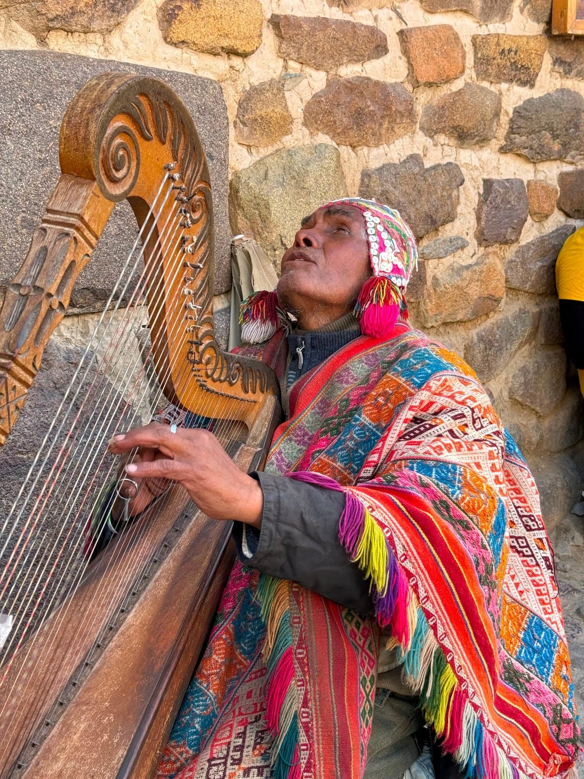 A blind Andean man in a bright woven poncho and knitted cap playing a carved wooden harp against a stone wall