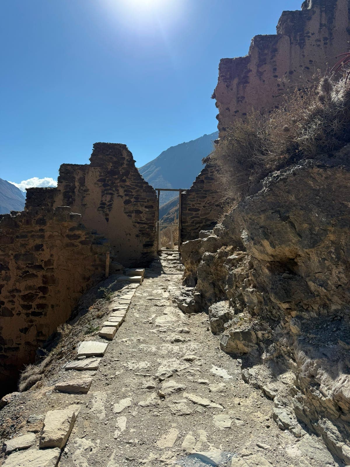 A stone pathway climbing between the ruined walls of Ollantaytambo toward a doorway, sun blazing over the mountains beyond
