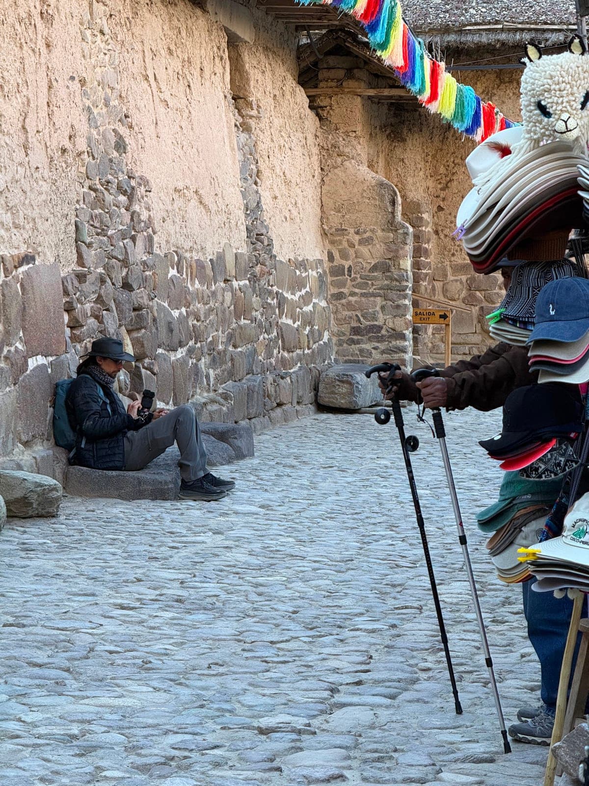 A cobblestone Inca street strung with rainbow bunting, a vendor's stall of knitted alpaca hats to one side and someone resting against the ancient wall