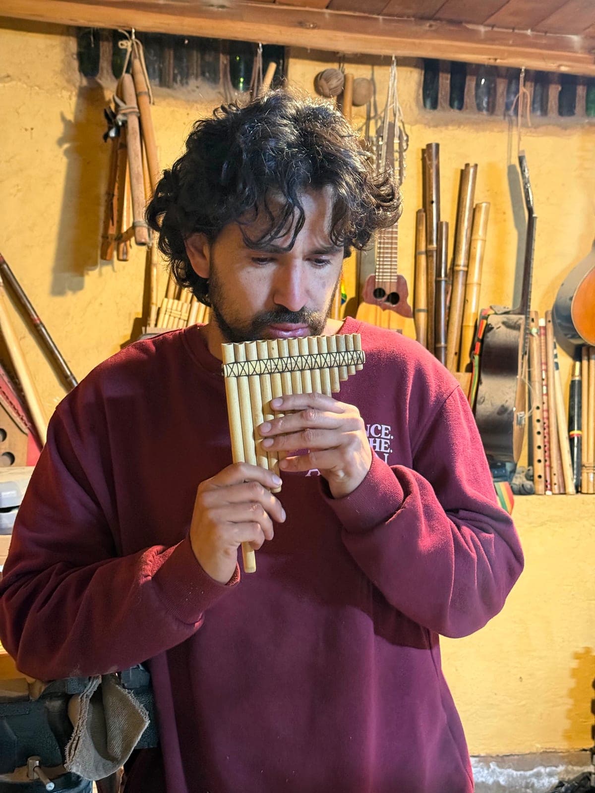 Omar in a maroon sweatshirt playing a set of bamboo panpipes in his workshop, flutes hanging on the wall behind him