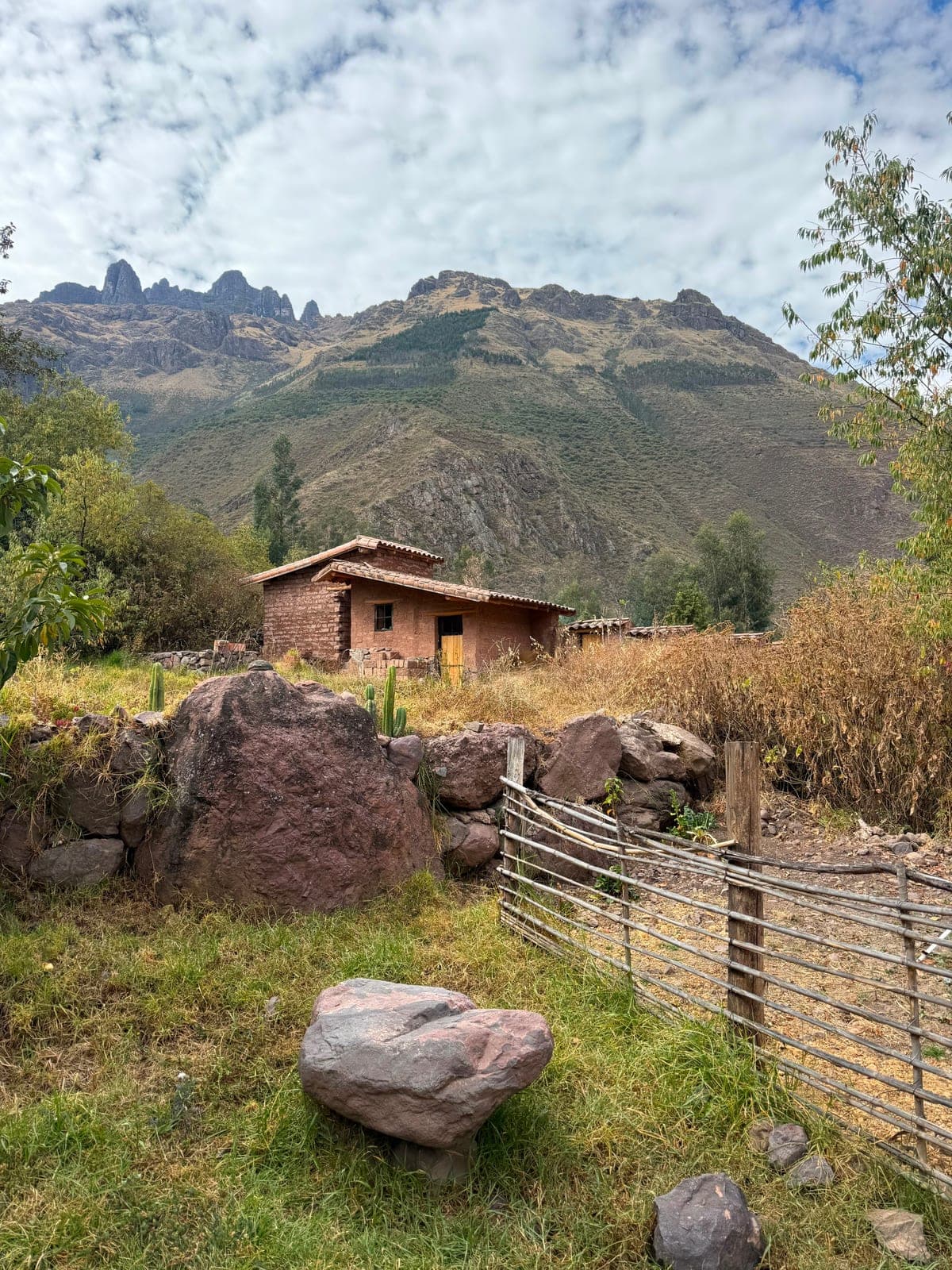 A small adobe house in the Sacred Valley set against jagged Andean peaks, a rustic reed-and-stone fence in the foreground