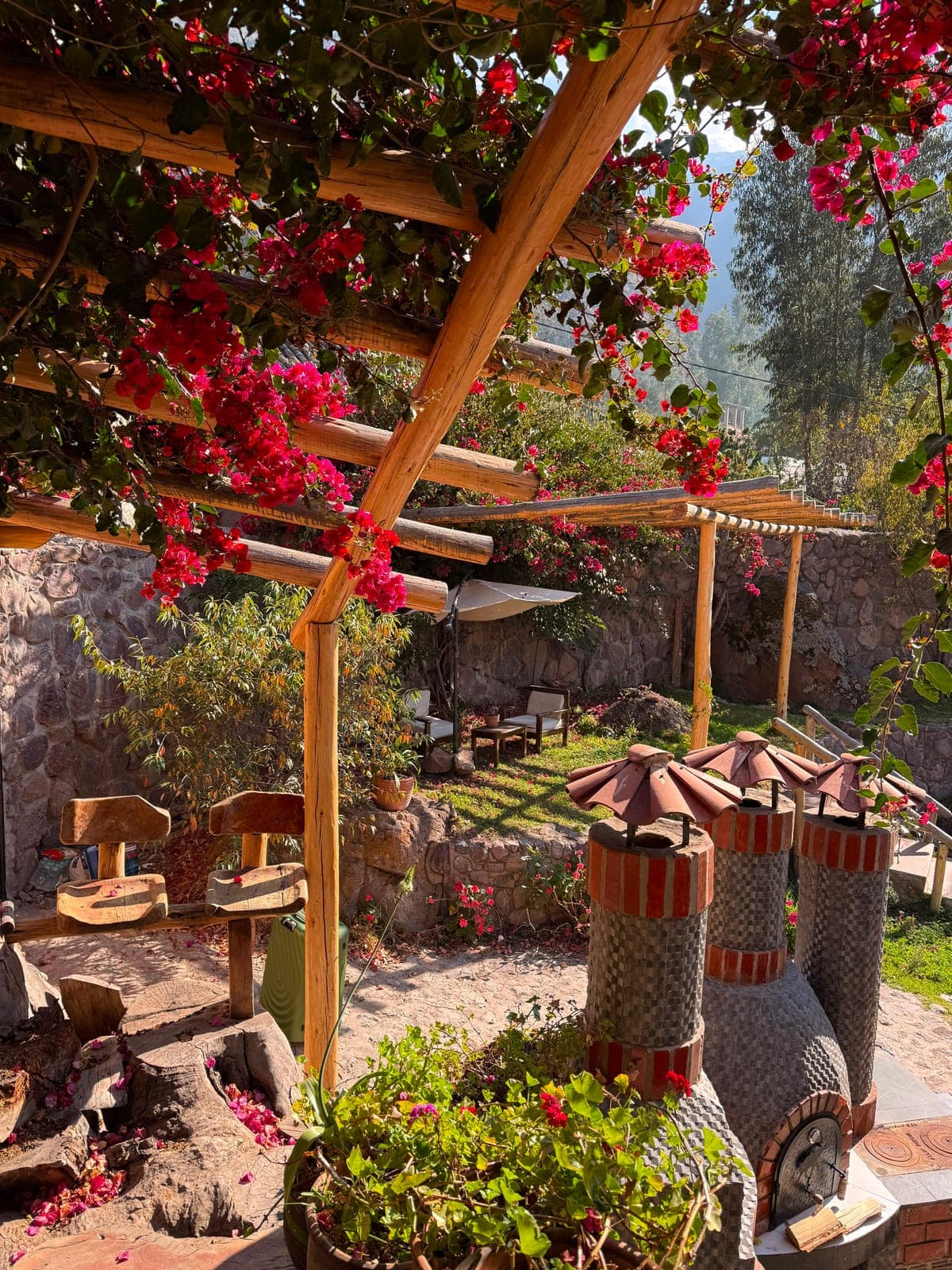 A sunny garden framed by bougainvillea over a wooden pergola, with clay bread ovens and mountains beyond