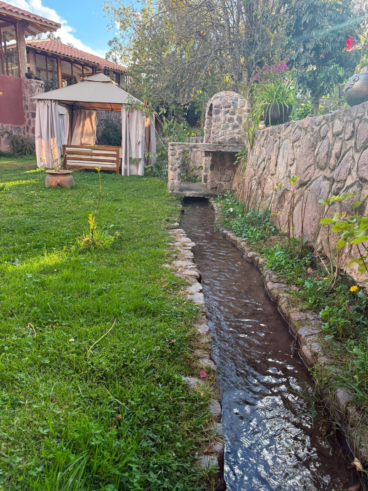 A clear mountain stream running through the grassy garden of the Airbnb, past a curtained cabana and a stone wall