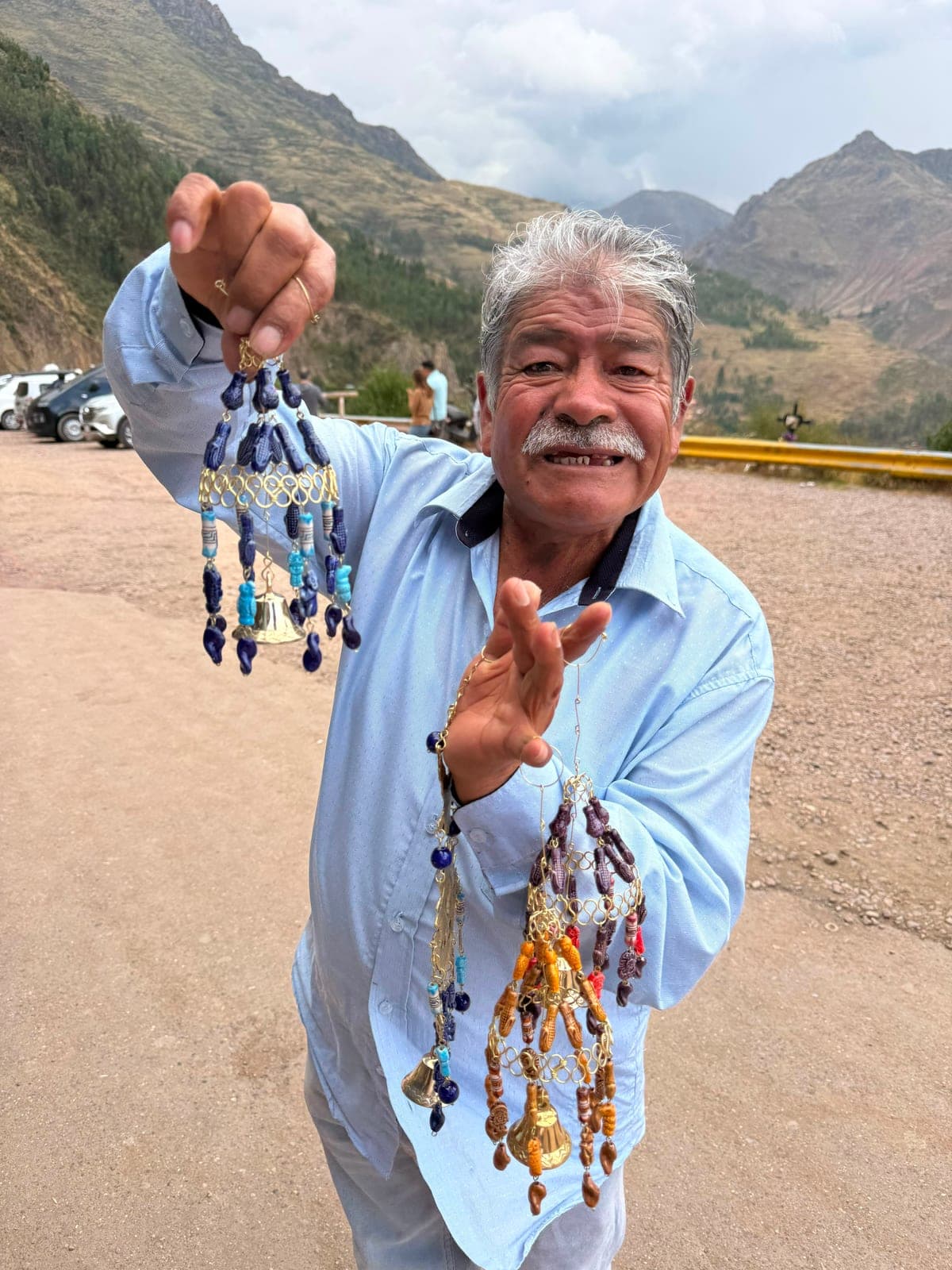 A smiling older man holding up handmade beaded ornaments and small brass bells at a roadside stop in the mountains