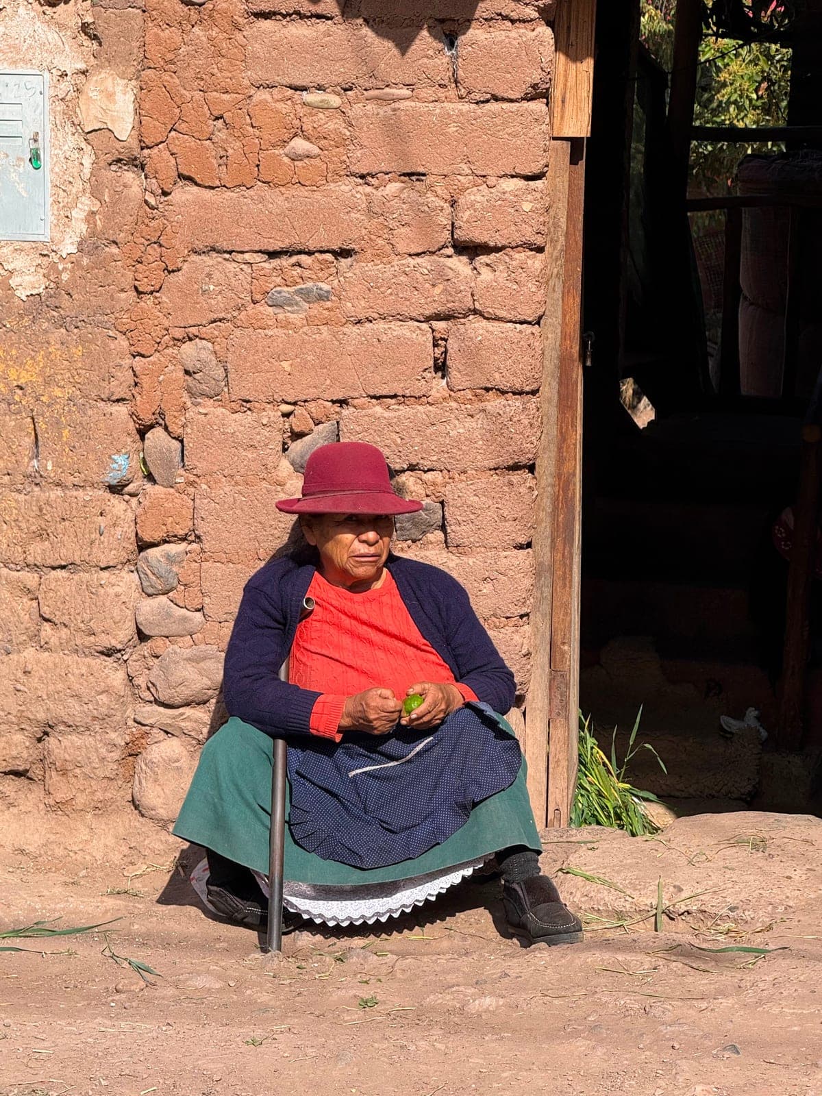 An elderly woman in a red hat resting on a low stool against an adobe wall
