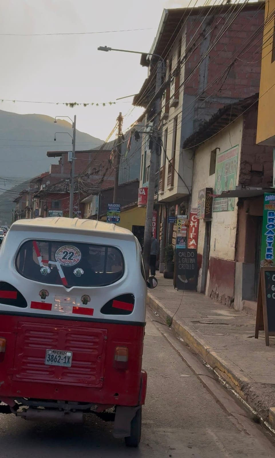 A red and white mototaxi driving down a narrow village street lined with small shops, mountains in the haze beyond