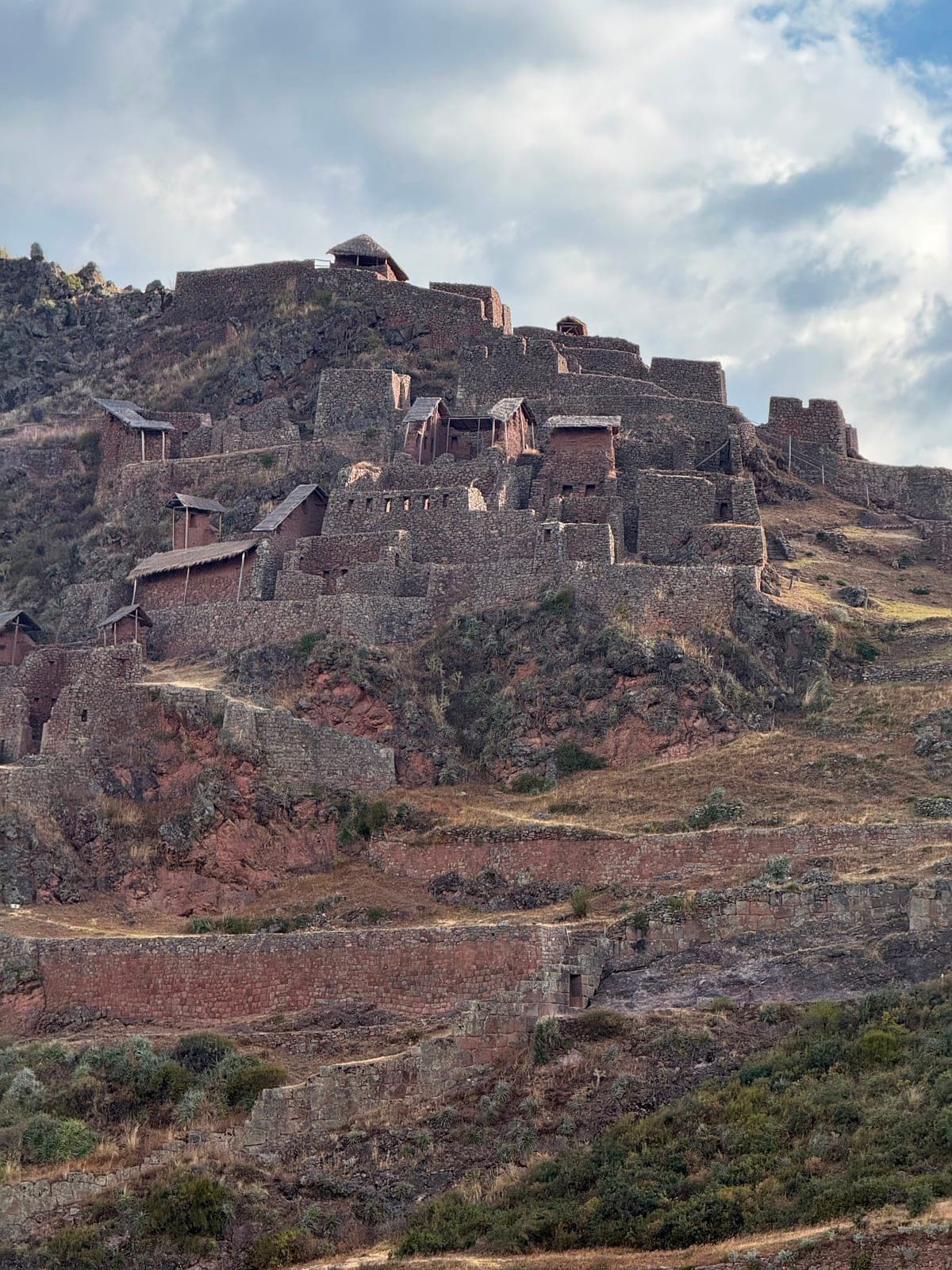 Tiers of Inca stone buildings and walls climbing a steep hillside under a cloudy sky