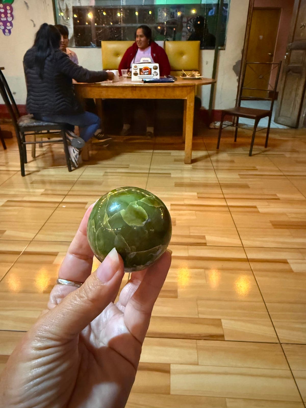 A hand holding a polished green crystal sphere inside a small local café, women seated at a table behind
