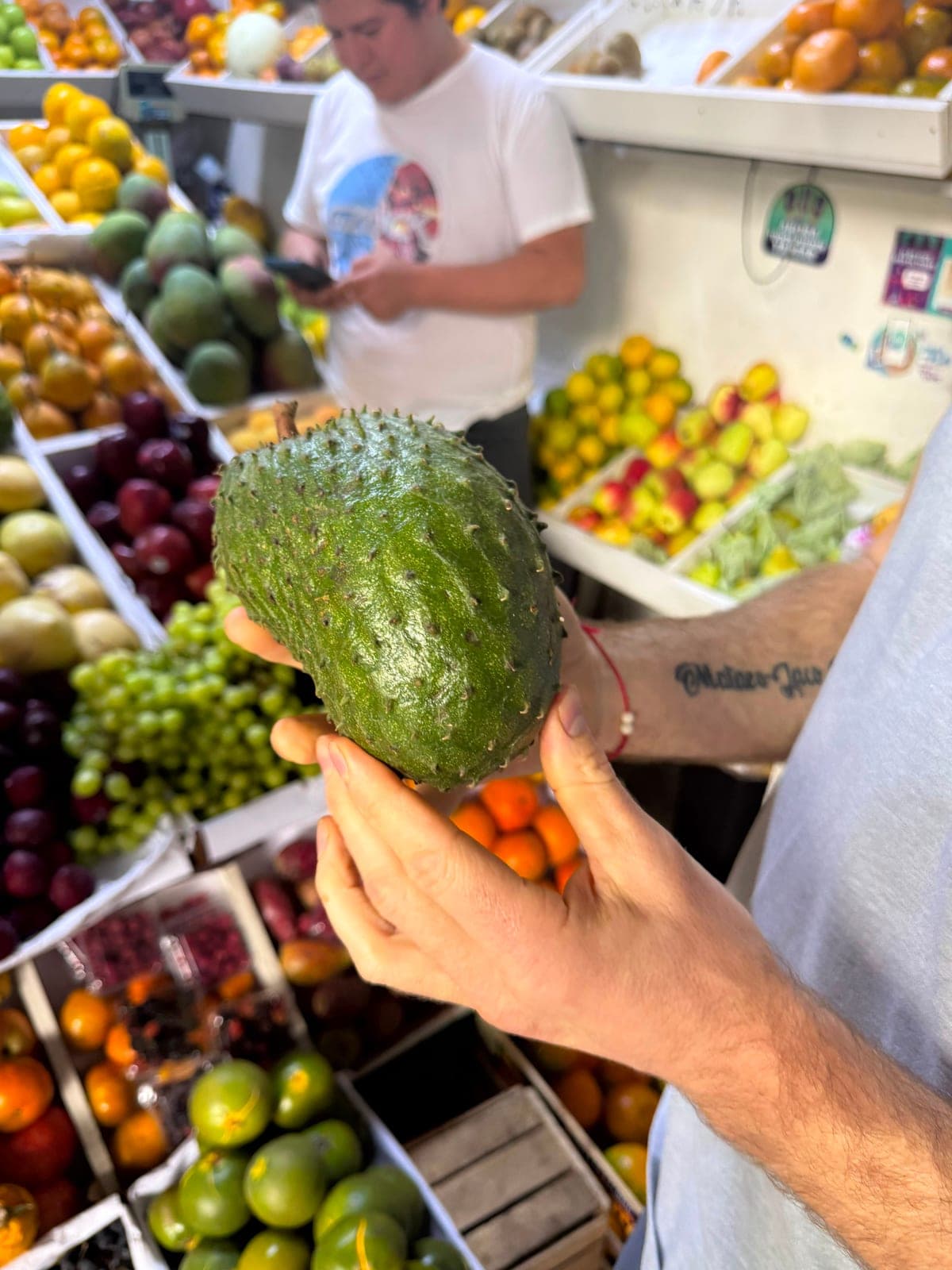 A hand holding a green spiny soursop fruit at the fruit market, stalls of produce behind