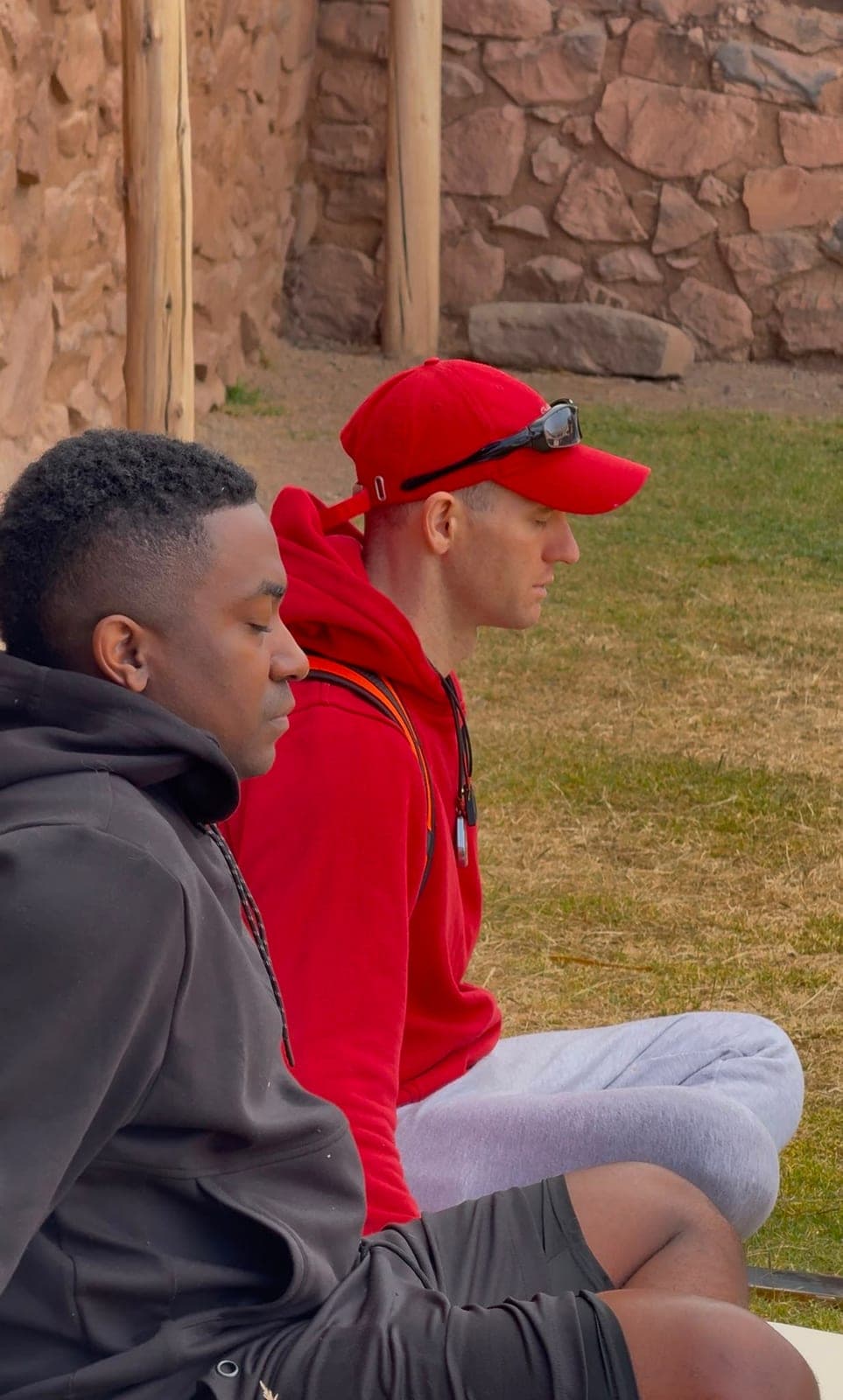 Two men sitting cross-legged with eyes closed, meditating on the grass among the stone ruins
