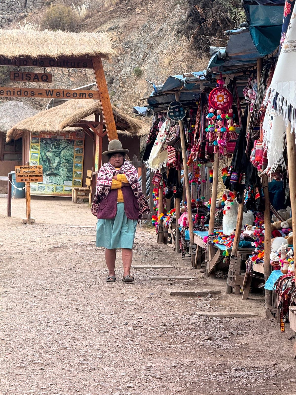 A wooden ‘PISAQ — Welcome’ gateway at the site entrance, a local woman and rows of colorful craft stalls alongside