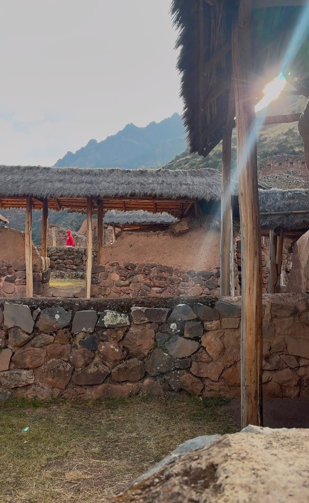 Restored Inca stone buildings with thatched roofs among the ruins, a figure in red in the distance and mountains behind