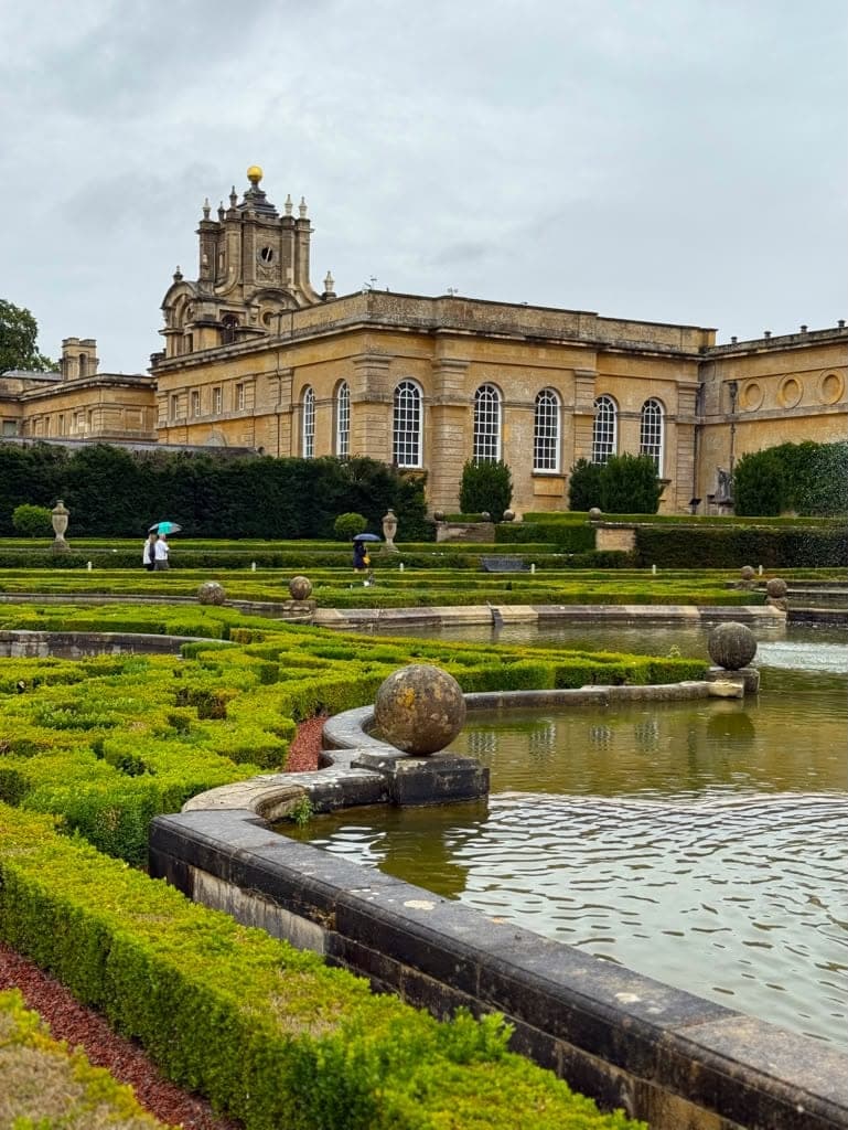 A formal water garden below a grand stone house, low box hedges and stone spheres around still pools, two people crossing the terrace under umbrellas in the rain