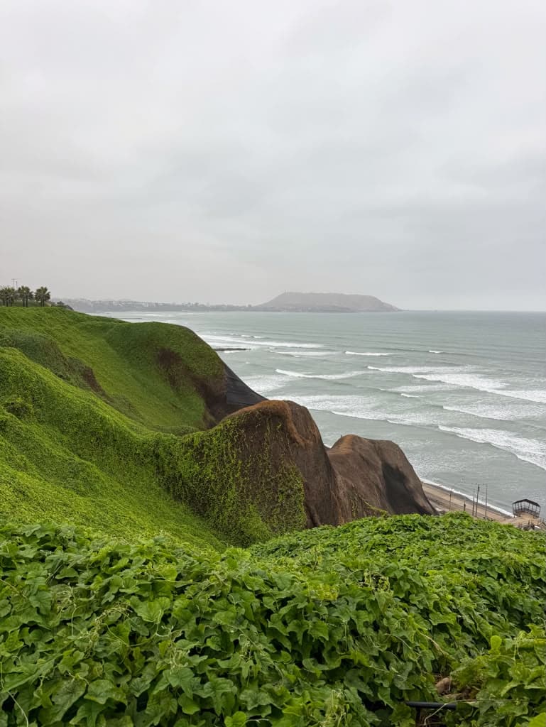 Green cliffs of Lima's Costa Verde dropping to a grey Pacific lined with breaking waves