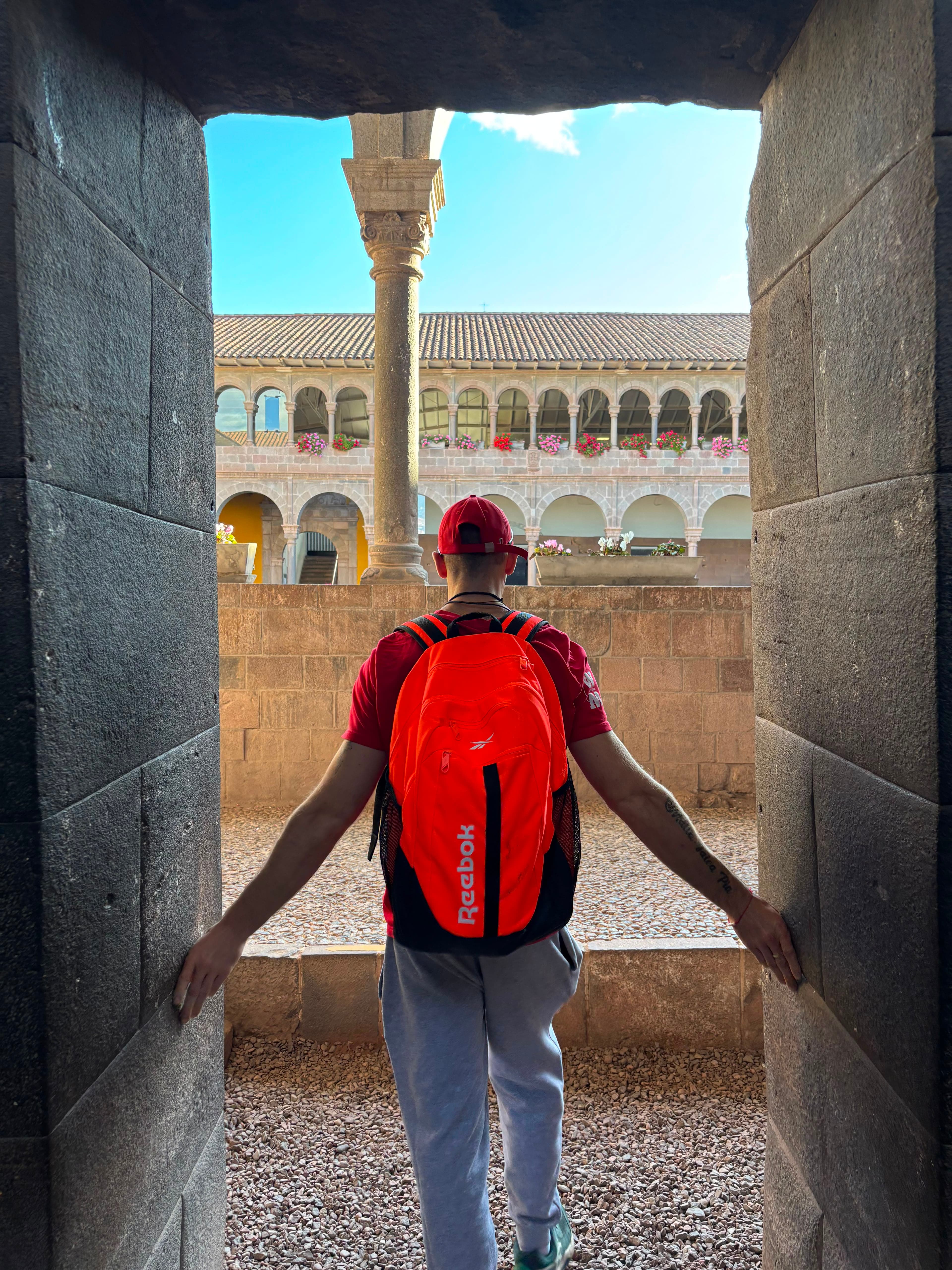 Jacob seen from behind wearing a bright orange Reebok backpack, walking through an Inca stone doorway in Cusco