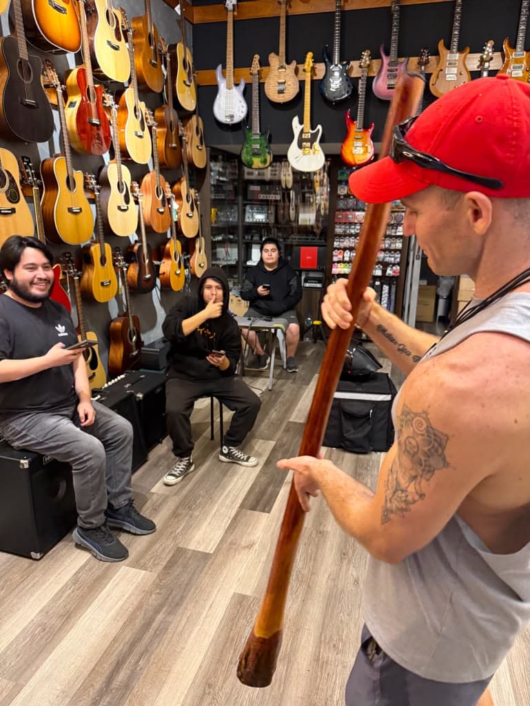 Jacob playing a long wooden yidaki inside a guitar shop while a few young men sit and watch, smiling