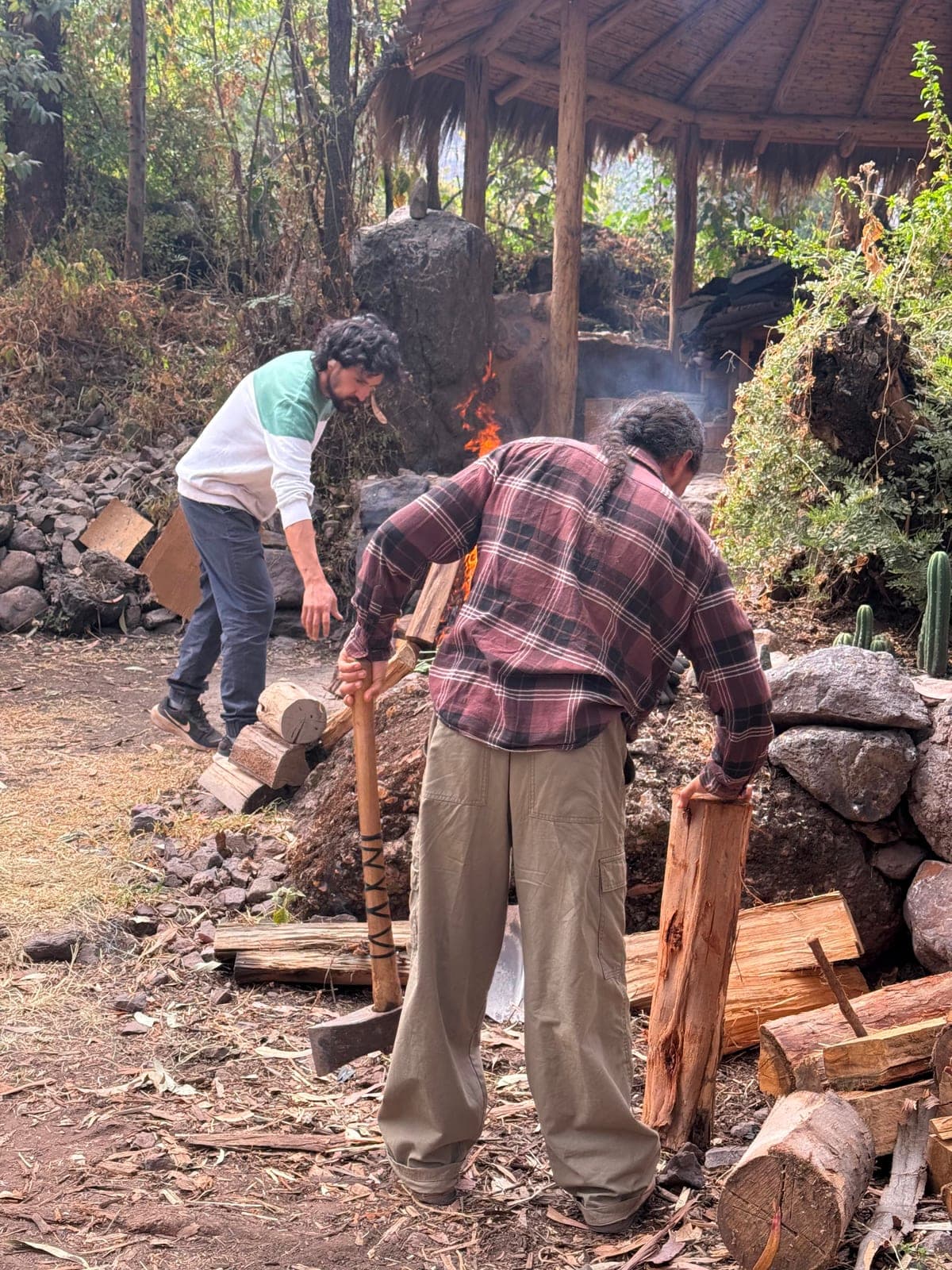 Two men splitting and stacking firewood beside an open fire where the ceremony stones are heated